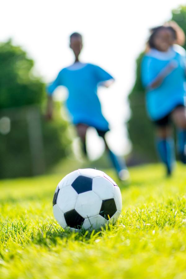 Kids play soccer in the background. A soccer ball is in focus in the foreground.